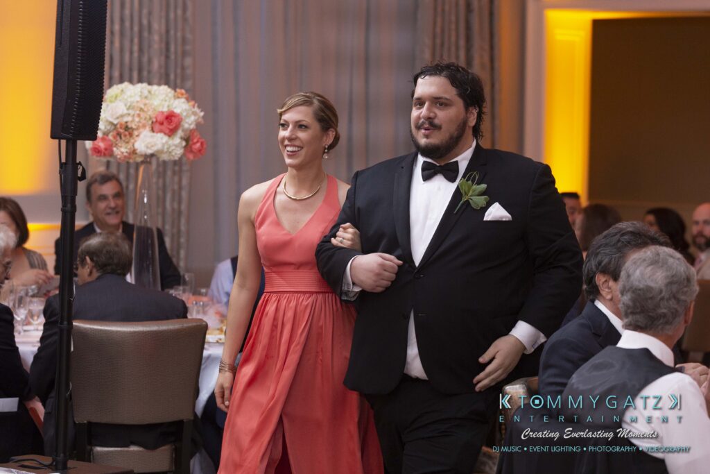 A woman in an orange dress and a man in a black tuxedo walk arm in arm in a formal event setting with seated guests.