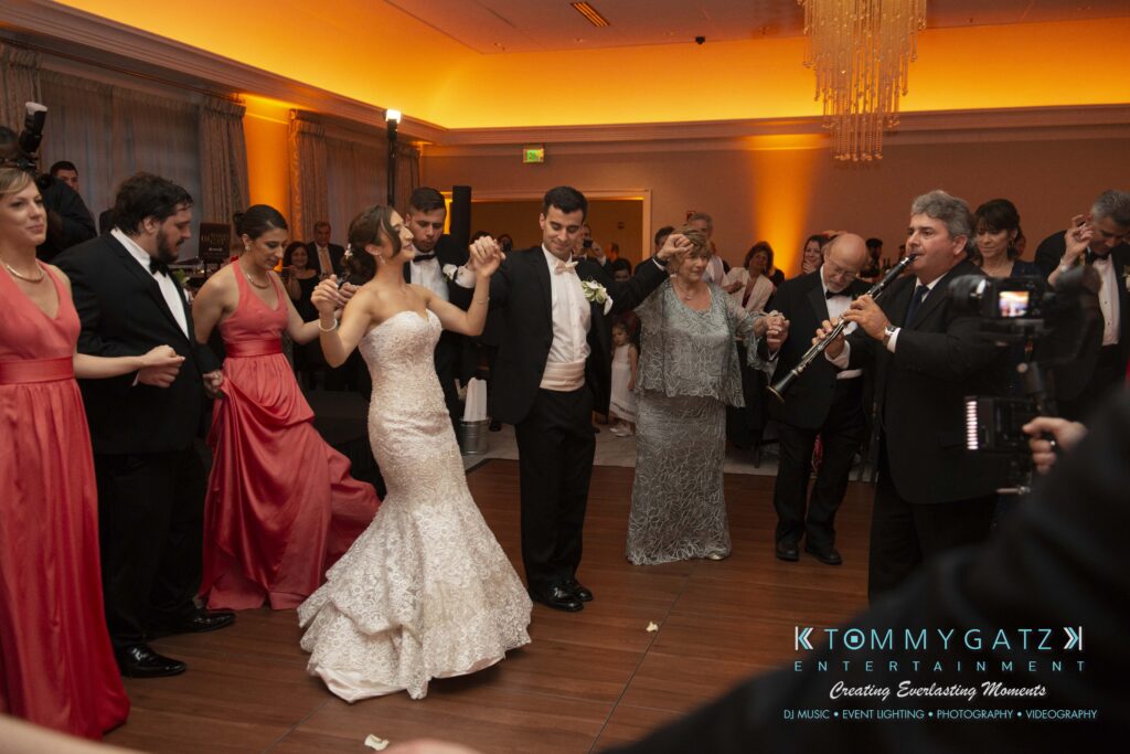 Bride and groom dance in a circle with guests at a formal event. People in elegant attire, some playing musical instruments, gather on a wooden dance floor.
