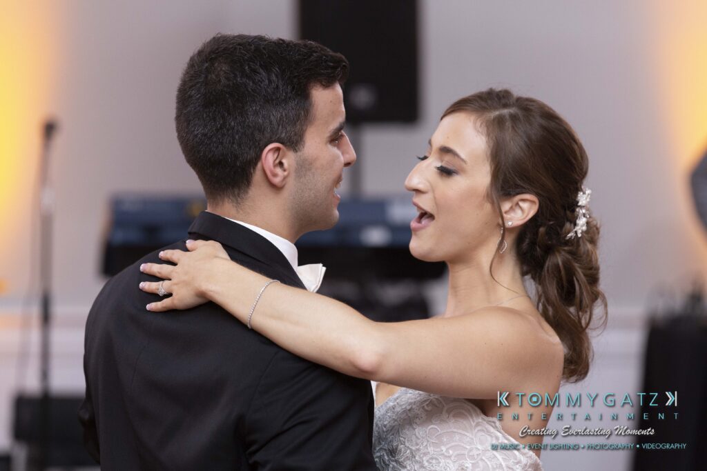 A couple dances together at a formal event, smiling and looking at each other. The background features blurred lights and equipment.