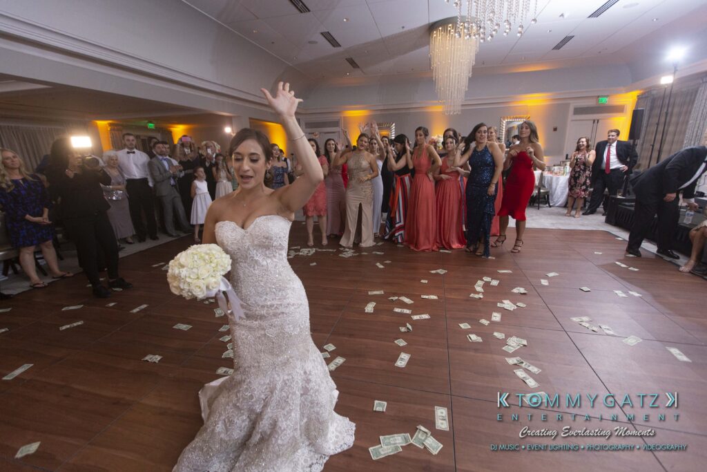 Bride in a wedding dress dances holding a bouquet on a money-covered floor, with guests clapping and cheering in the background.