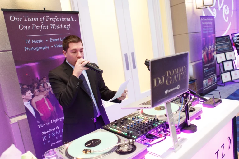 A DJ in a suit speaks into a microphone at an event booth with DJ equipment, standing beside a promotional wedding services banner.